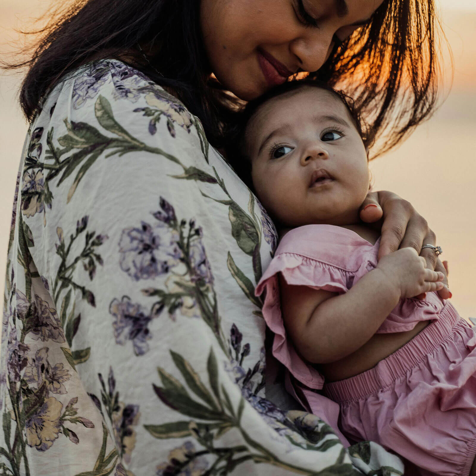 A mother dressed in floral attire holding her baby close, gazing at the baby in soft natural light.